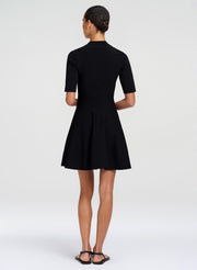 A woman viewed from the back wearing a black short-sleeve fit-and-flare dress with a high mock neck and pleated skirt, paired with black flat sandals, against a light gray studio background.