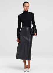 A model wearing a black long-sleeve mock neck top, a black leather midi skirt, and white pointed-toe heels, holding a black clutch against a light gray studio background.