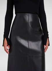 A close-up of a model wearing a black leather midi skirt with a front seam and a black ribbed long-sleeve top with split cuffs, against a light gray studio background.