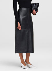 A model wearing a black leather midi skirt, a black long-sleeve top, white pointed-toe flats, and holding a black clutch bag against a light gray studio background.
