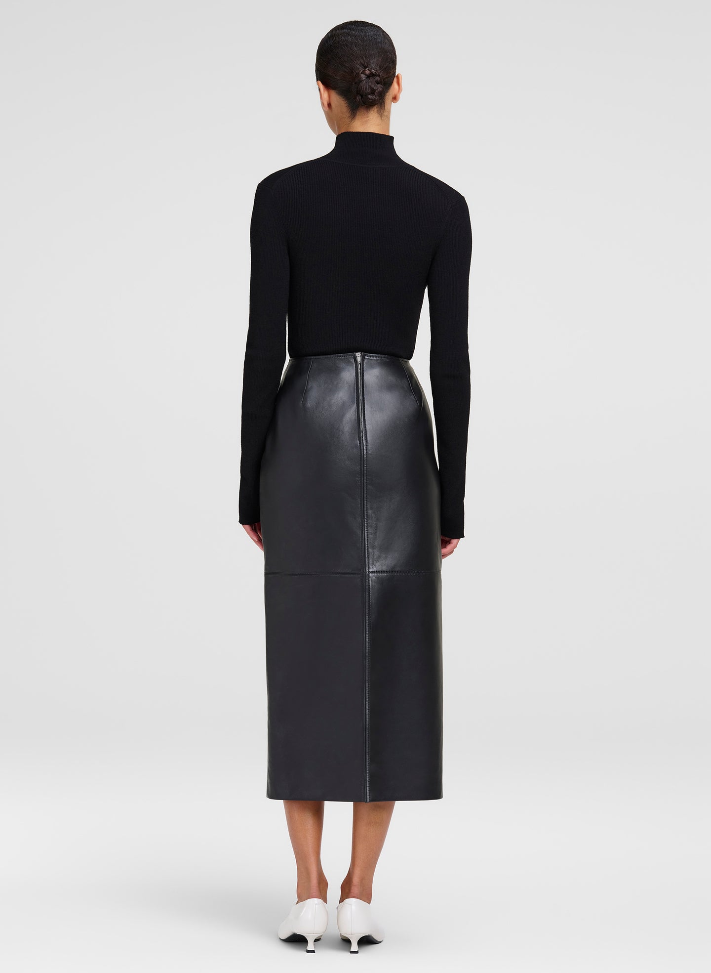 A model shown from the back wearing a black long-sleeve mock-neck top and a black leather midi skirt, paired with white pointed-toe heels, against a light gray studio background.