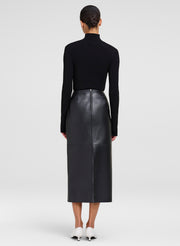 A model shown from the back wearing a black long-sleeve mock-neck top and a black leather midi skirt, paired with white pointed-toe heels, against a light gray studio background.