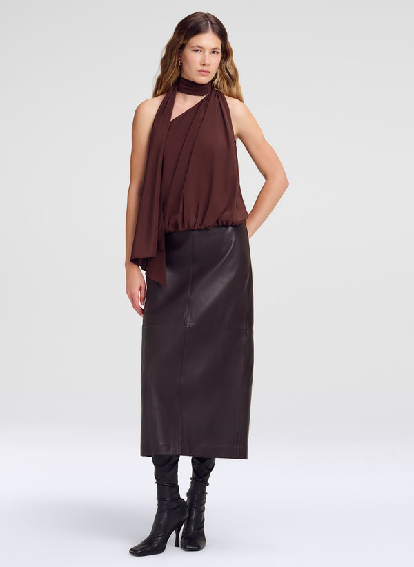 A model wearing a dark brown halter-neck draped top, a dark brown leather midi skirt, and black heeled boots, photographed against a light gray studio background.