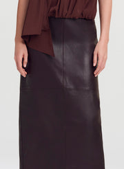 A close-up of a model wearing a dark brown leather midi skirt paired with a matching dark brown draped top, shown against a light gray studio background.