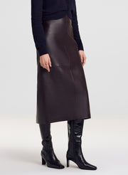 A model wearing a dark brown leather midi skirt, a navy blue long-sleeve top, and black leather knee-high boots, shown from the waist down on a light gray studio background.
