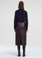 A model viewed from the back wearing a dark navy long-sleeve knit top, a dark brown leather midi skirt with a back slit, and black knee-high boots, against a light gray studio background.