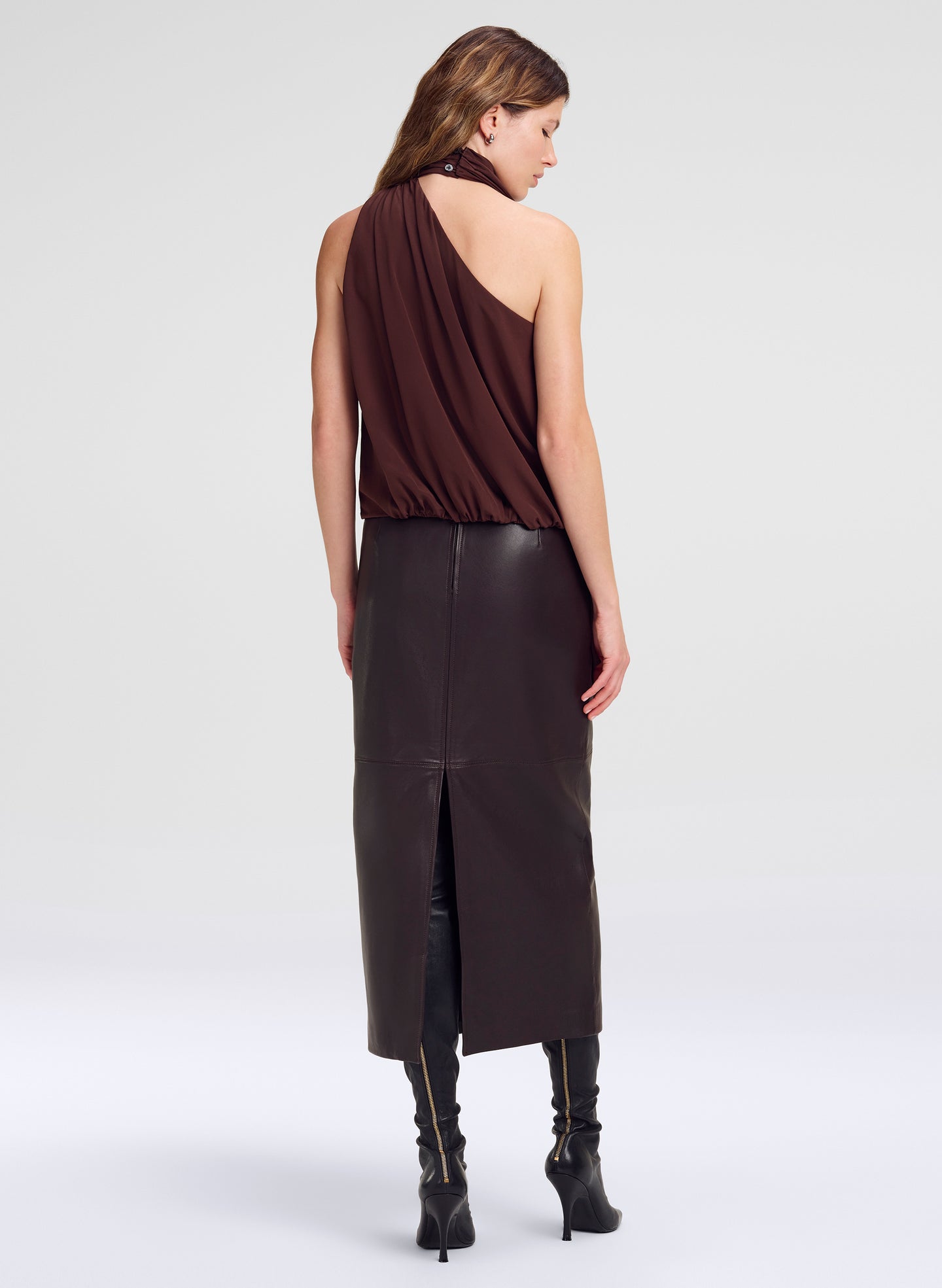 A model is shown from the back wearing a dark brown draped one-shoulder top, a brown leather midi skirt with a front slit, and black heeled boots, set against a plain light gray studio background.