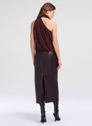 A model is shown from the back wearing a dark brown draped one-shoulder top, a brown leather midi skirt with a front slit, and black heeled boots, set against a plain light gray studio background.