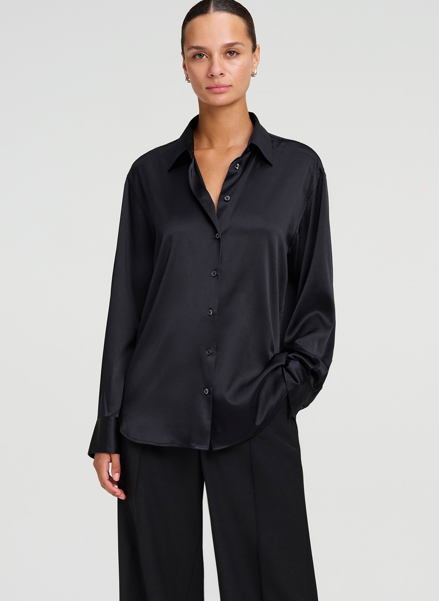 A woman modeling a black long-sleeve button-up shirt with a silk satin sheen, paired with black wide-leg trousers, against a light gray studio background.