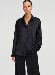 A woman modeling a black long-sleeve button-up shirt with a silk satin sheen, paired with black wide-leg trousers, against a light gray studio background.