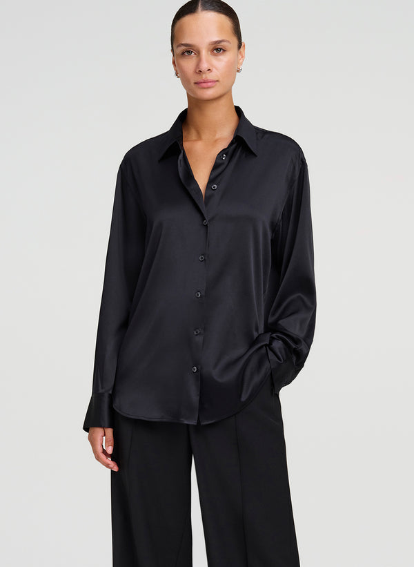 A woman modeling a black long-sleeve button-up shirt with a silk satin sheen, paired with black wide-leg trousers, against a light gray studio background.
