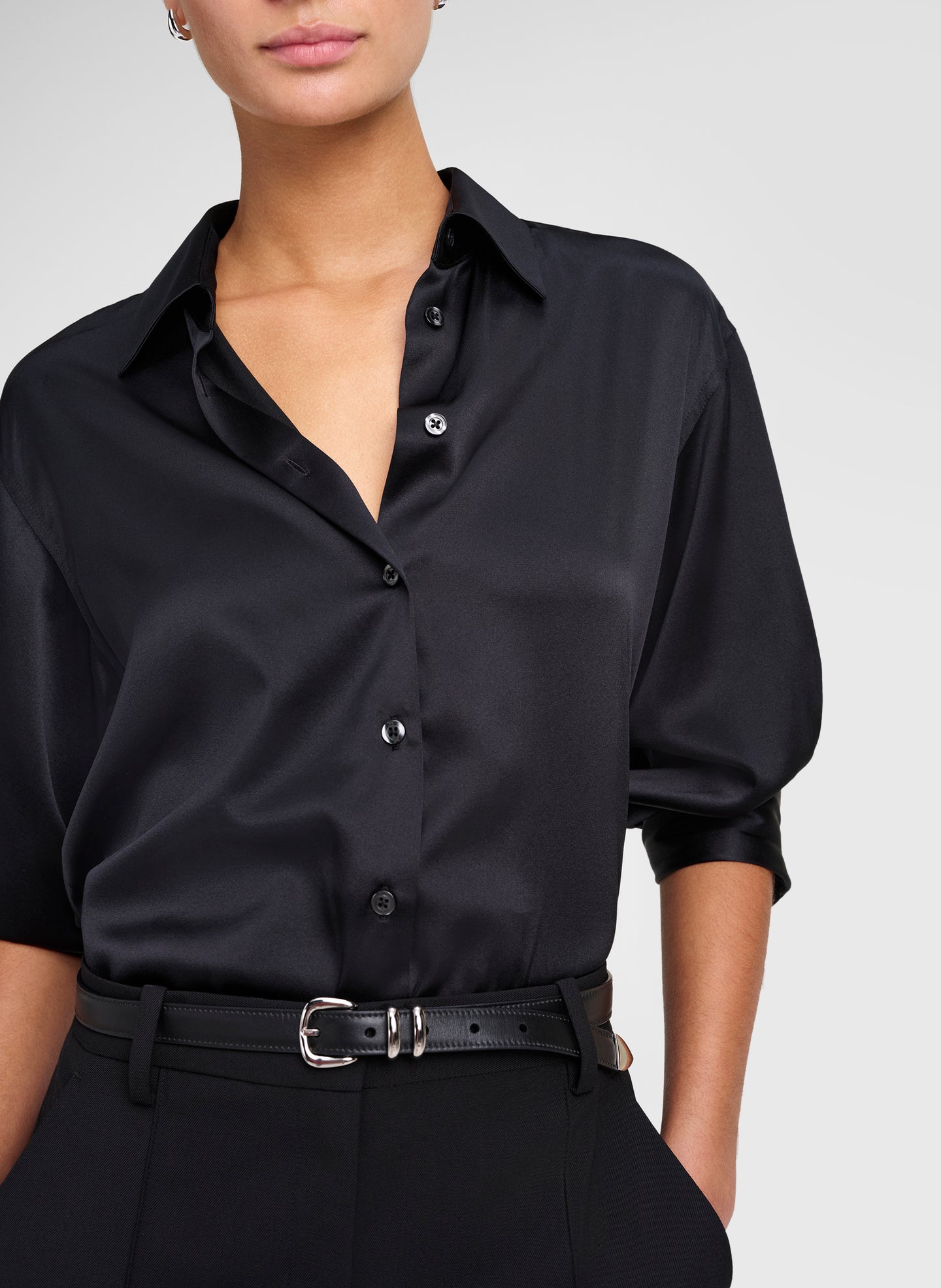 A close-up of a model wearing a black satin button-down shirt with three-quarter sleeves, tucked into black trousers and secured with a black leather belt featuring a silver buckle.