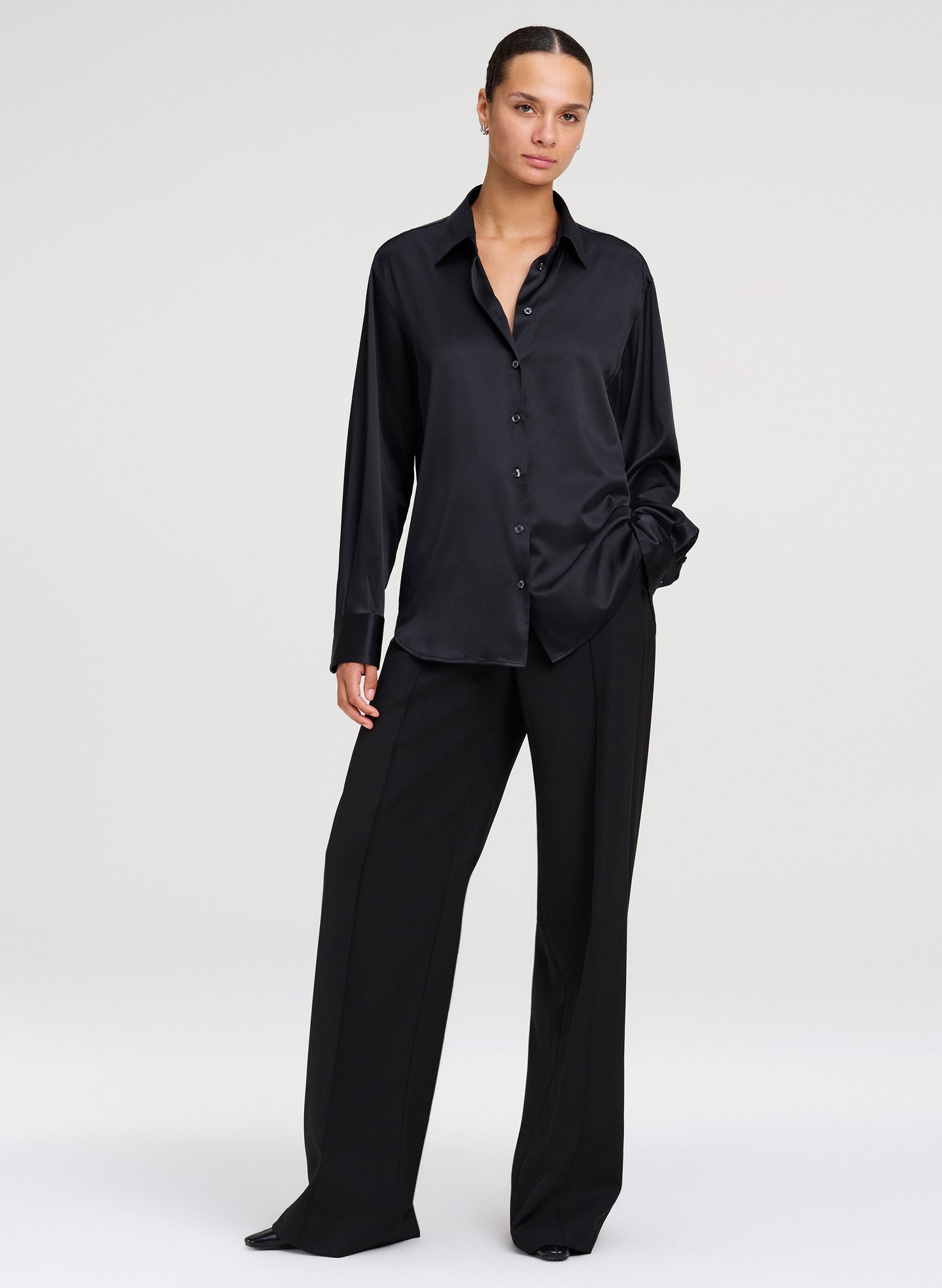 A model wearing a black long-sleeve button-up shirt and matching black wide-leg trousers, standing against a light gray studio background.