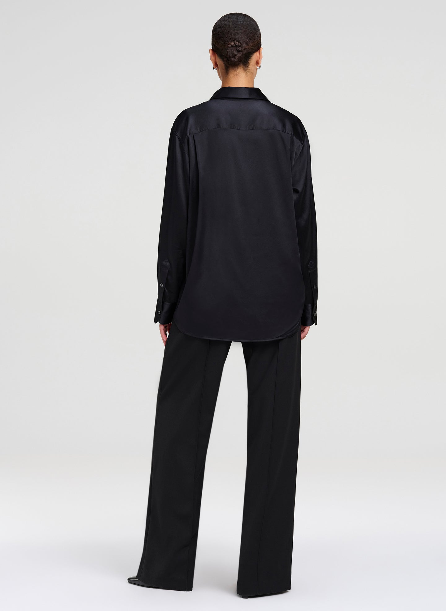 A model viewed from the back wearing a black long-sleeve satin shirt and matching black wide-leg trousers against a light gray studio background.