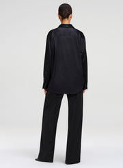 A model viewed from the back wearing a black long-sleeve satin shirt and matching black wide-leg trousers against a light gray studio background.