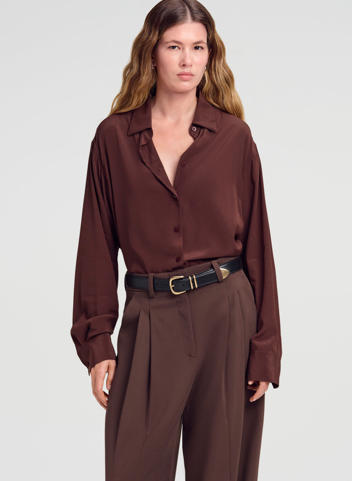 A woman modeling a dark brown long-sleeve button-up shirt with matching brown trousers and a black belt, photographed against a light gray studio background.