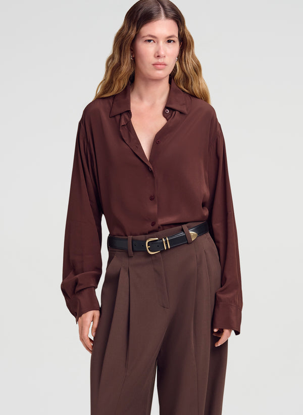 A woman modeling a dark brown long-sleeve button-up shirt with matching brown trousers and a black belt, photographed against a light gray studio background.