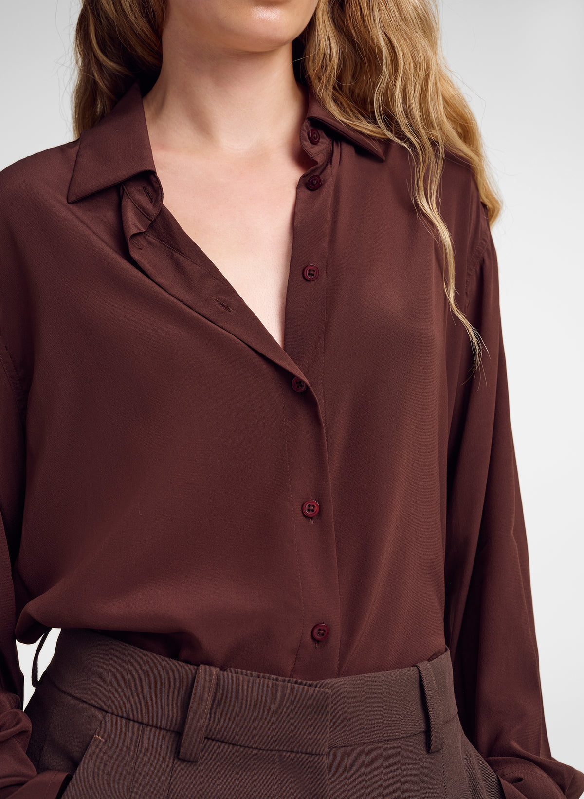 A close-up of a woman wearing a long-sleeve button-up blouse in a deep chocolate brown color, paired with matching brown trousers.