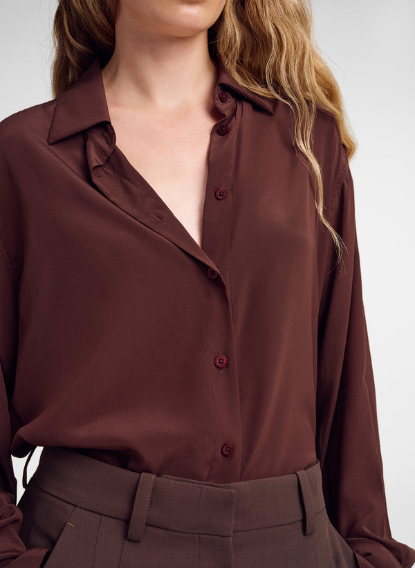 A close-up of a woman wearing a long-sleeve button-up blouse in a deep chocolate brown color, paired with matching brown trousers.