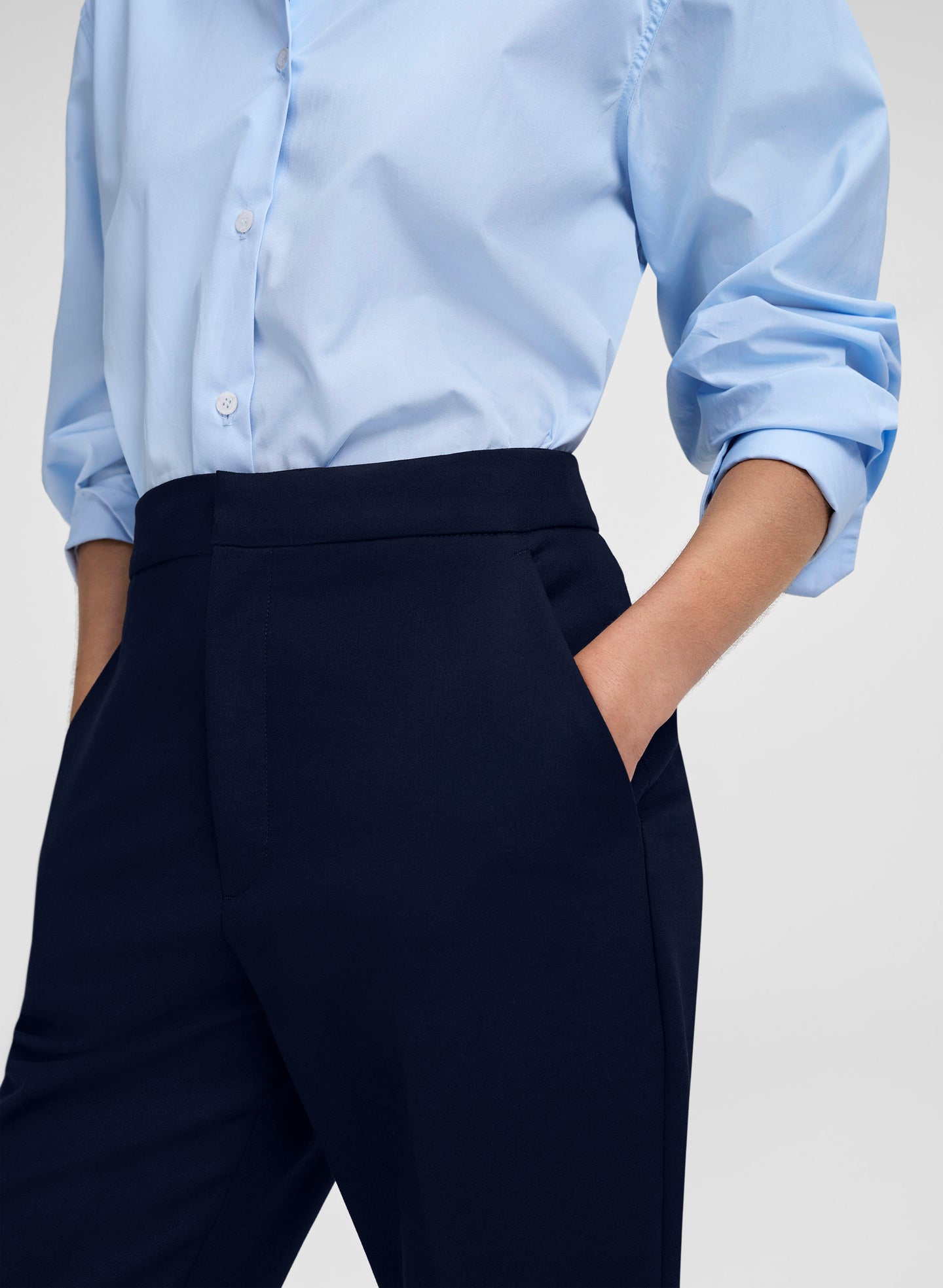 A close-up of a person wearing a light blue long-sleeve button-up shirt tucked into dark navy trousers, with one hand in a pocket, against a light gray studio background.