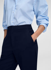 A close-up of a person wearing a light blue long-sleeve button-up shirt tucked into dark navy trousers, with one hand in a pocket, against a light gray studio background.