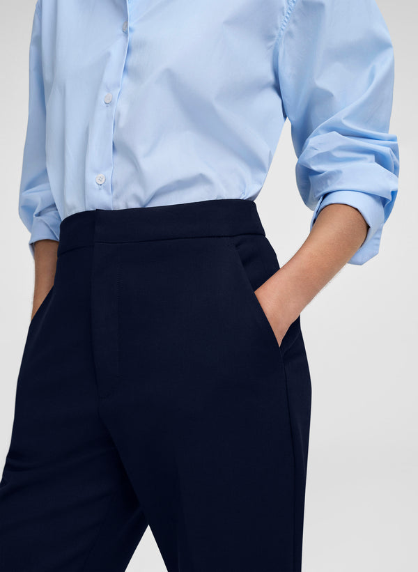 A close-up of a person wearing a light blue long-sleeve button-up shirt tucked into dark navy trousers, with one hand in a pocket, against a light gray studio background.