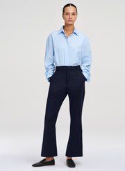 A model stands against a light gray studio background wearing a light blue long-sleeve button-up shirt, dark navy wide-leg trousers, and black loafers.