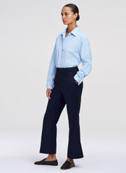 A woman modeling a light blue long-sleeve button-up shirt tucked into dark navy wide-leg trousers, paired with black slip-on loafers, against a plain light gray studio background.