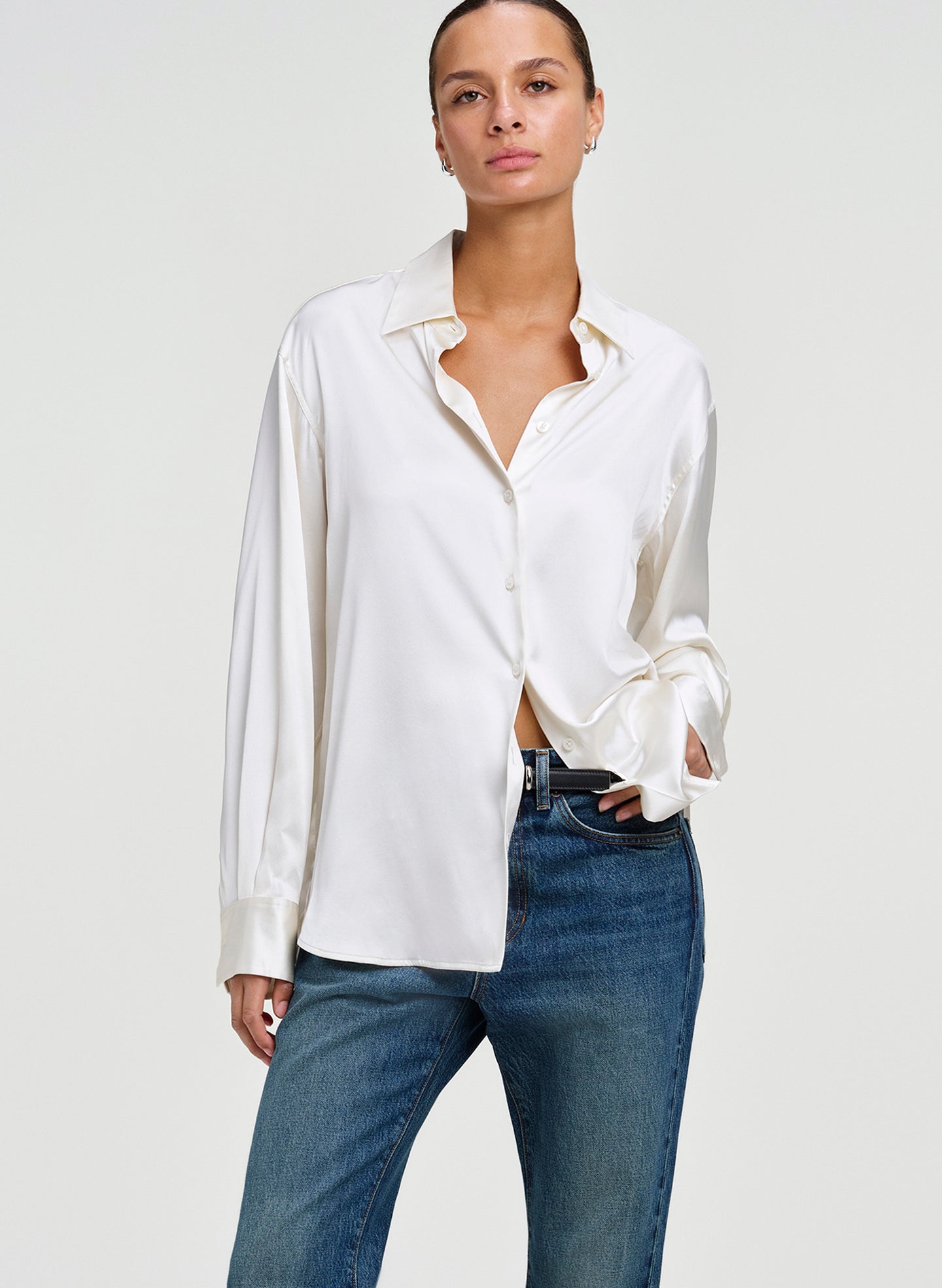 A woman modeling a white long-sleeve button-up shirt with a relaxed, slightly open collar and cuffs, paired with blue jeans, against a light gray studio background.