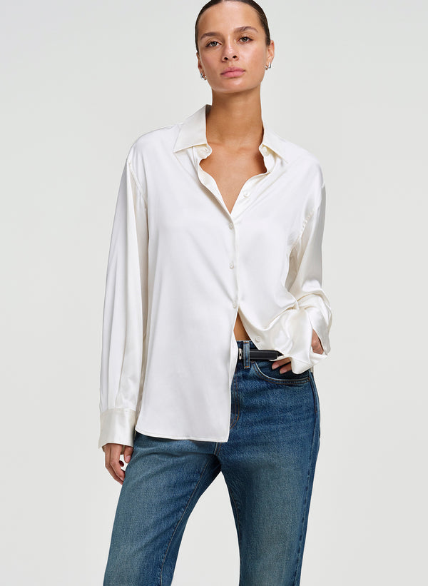 A woman modeling a white long-sleeve button-up shirt with a relaxed, slightly open collar and cuffs, paired with blue jeans, against a light gray studio background.