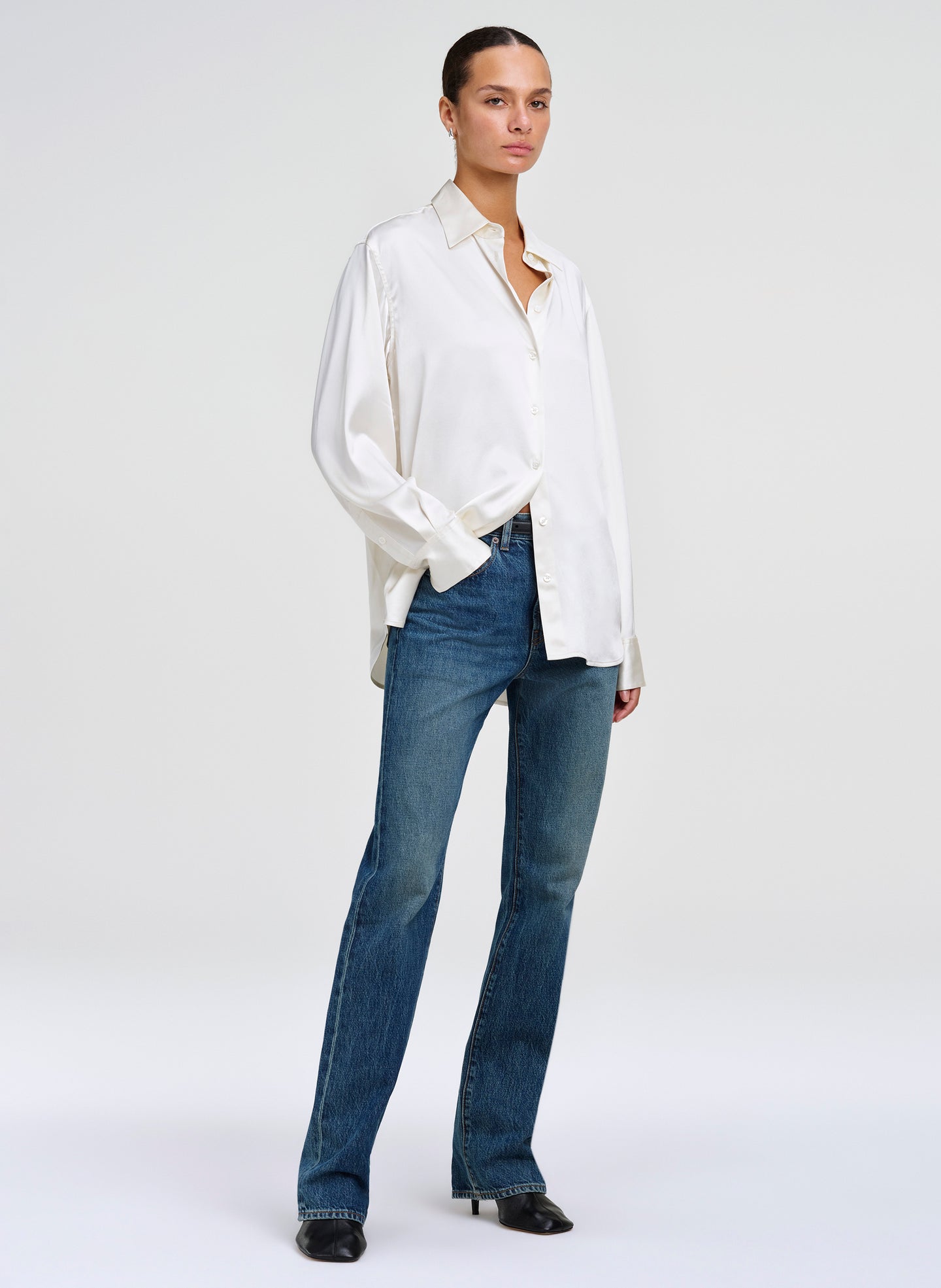 A model wearing a white long-sleeve button-down shirt and blue jeans, standing against a light gray studio background.