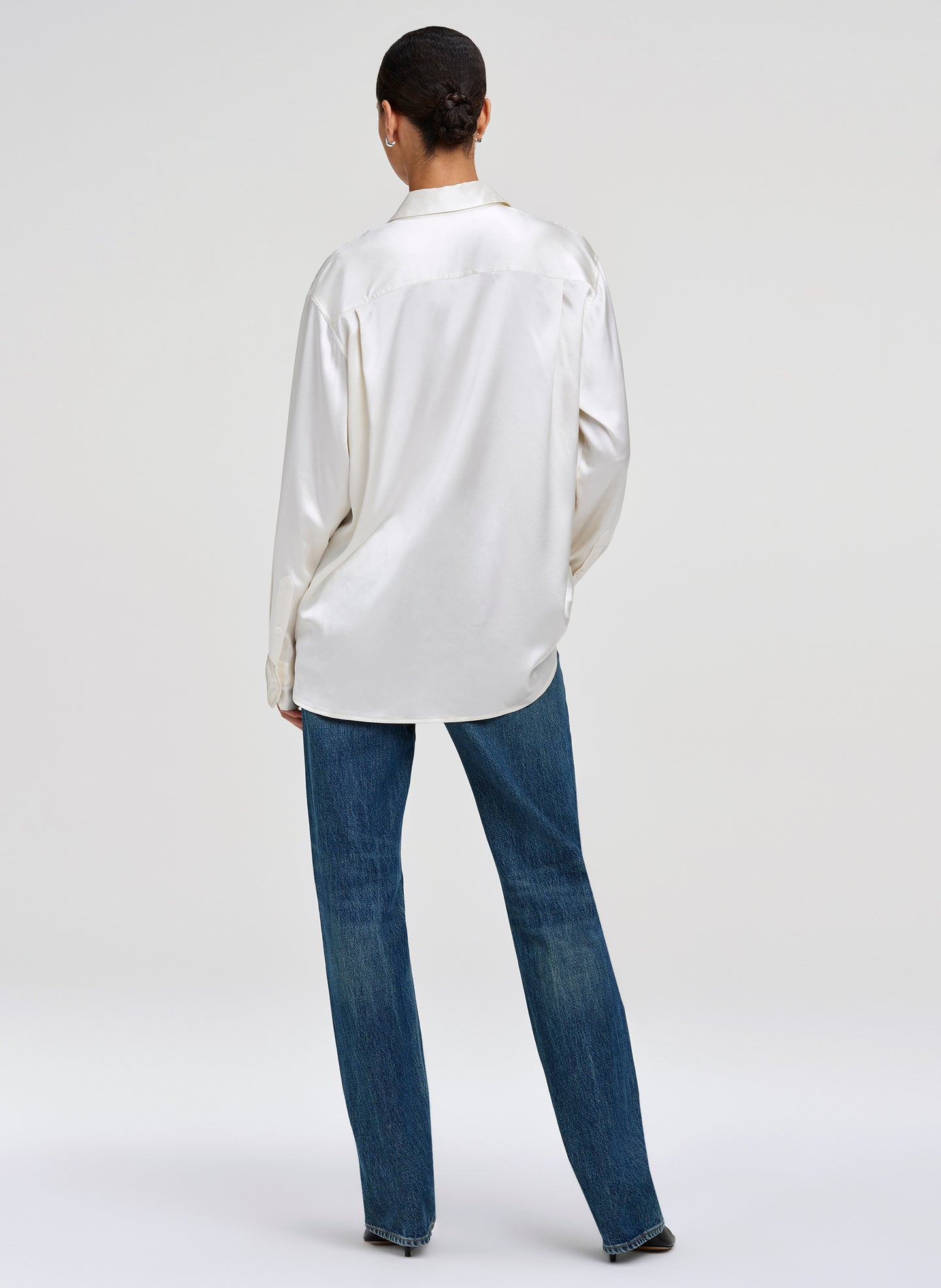 A model shown from the back wearing a white satin long-sleeve button-up shirt and blue jeans against a light gray studio background.