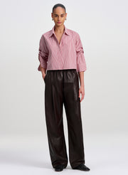 A model stands against a light gray studio background wearing a cropped maroon striped button-up shirt with rolled sleeves and dark brown wide-leg pants.