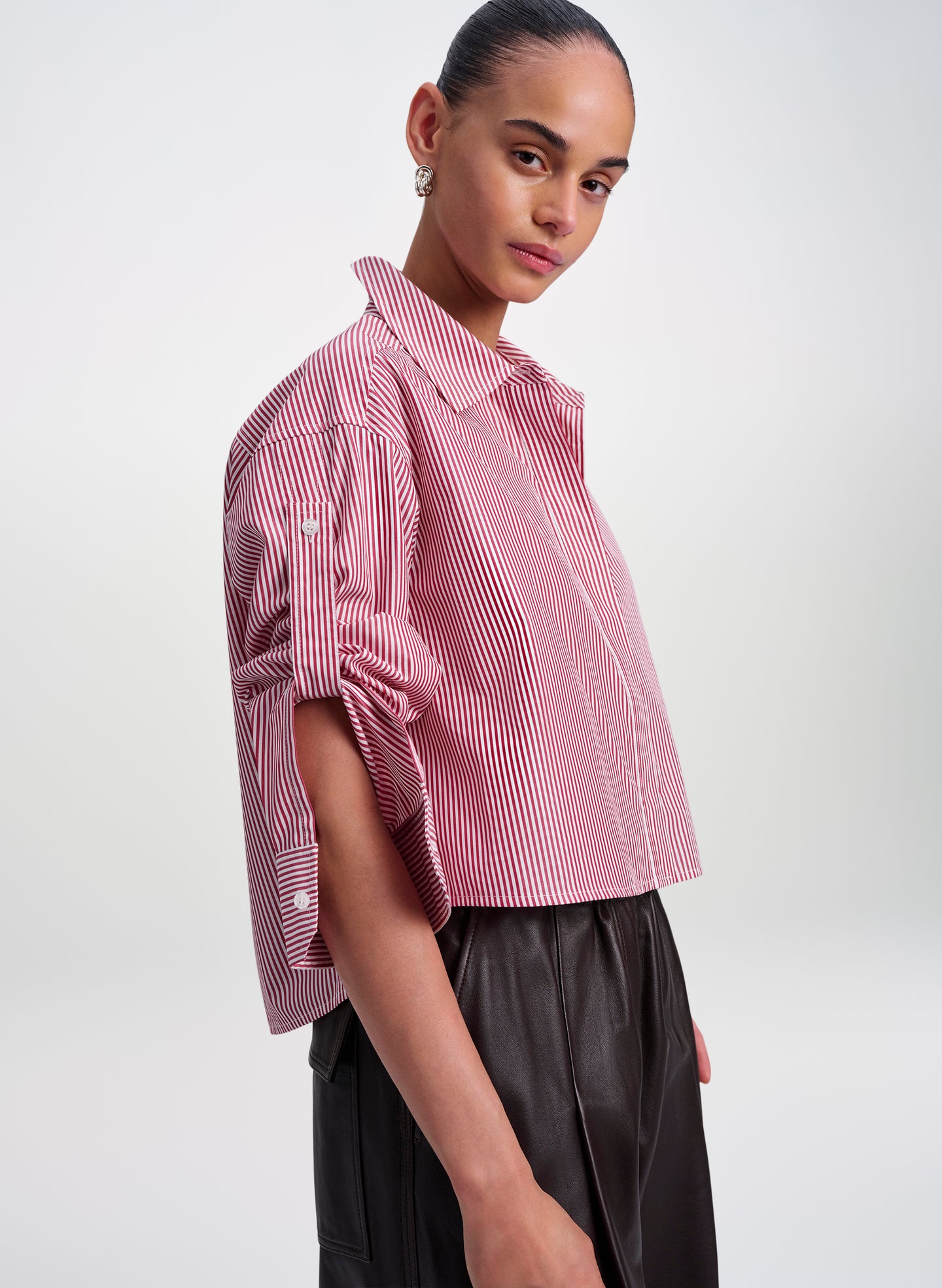 A model wearing a cropped red and white vertically striped button-up shirt with rolled sleeves, paired with dark pleated trousers, against a light gray studio background.