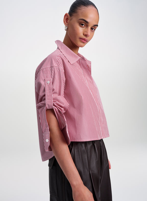 A model wearing a cropped red and white vertically striped button-up shirt with rolled sleeves, paired with dark pleated trousers, against a light gray studio background.
