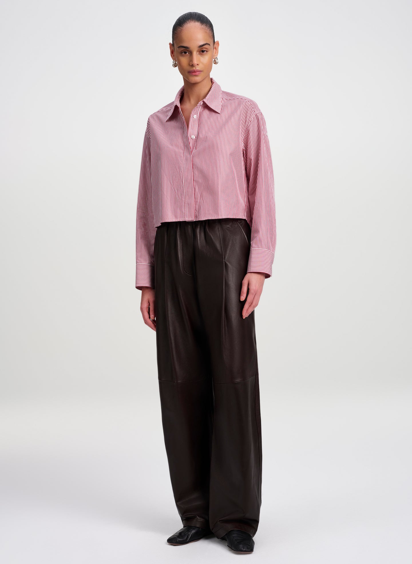 A model stands against a light gray studio background wearing a cropped maroon striped button-up shirt with subtle vertical texture and dark brown wide-leg pants.