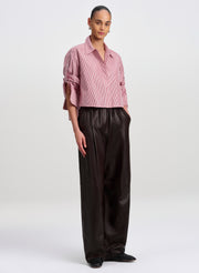 A model stands against a light gray studio background wearing a cropped striped button-up shirt with rolled sleeves and dark brown wide-leg satin trousers.