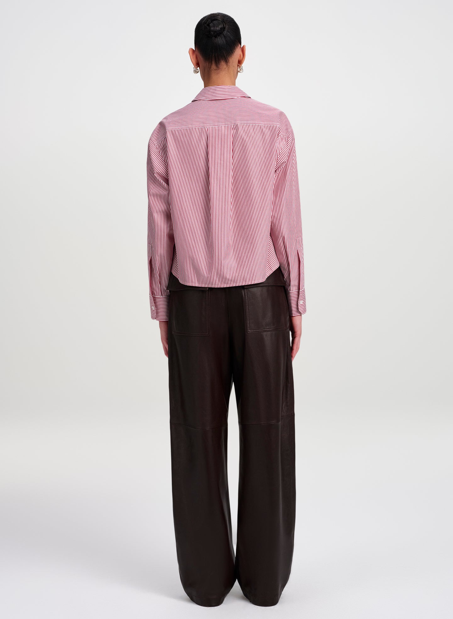 A model viewed from the back wearing a striped red and white long-sleeve button-up shirt and dark brown wide-leg trousers, set against a plain light gray studio background.