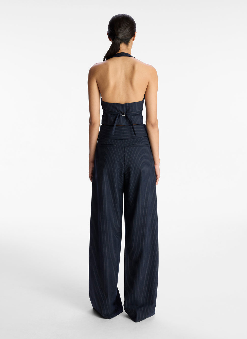 back view of  view of woman wearing navy blue pinstripe suit comprised of vest and fold over wide leg pants