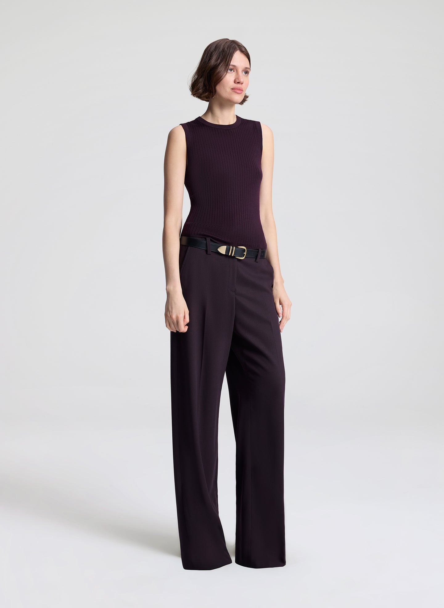 Woman wearing a purple sleeveless top and purple pants, holding a matching handbag against a light background