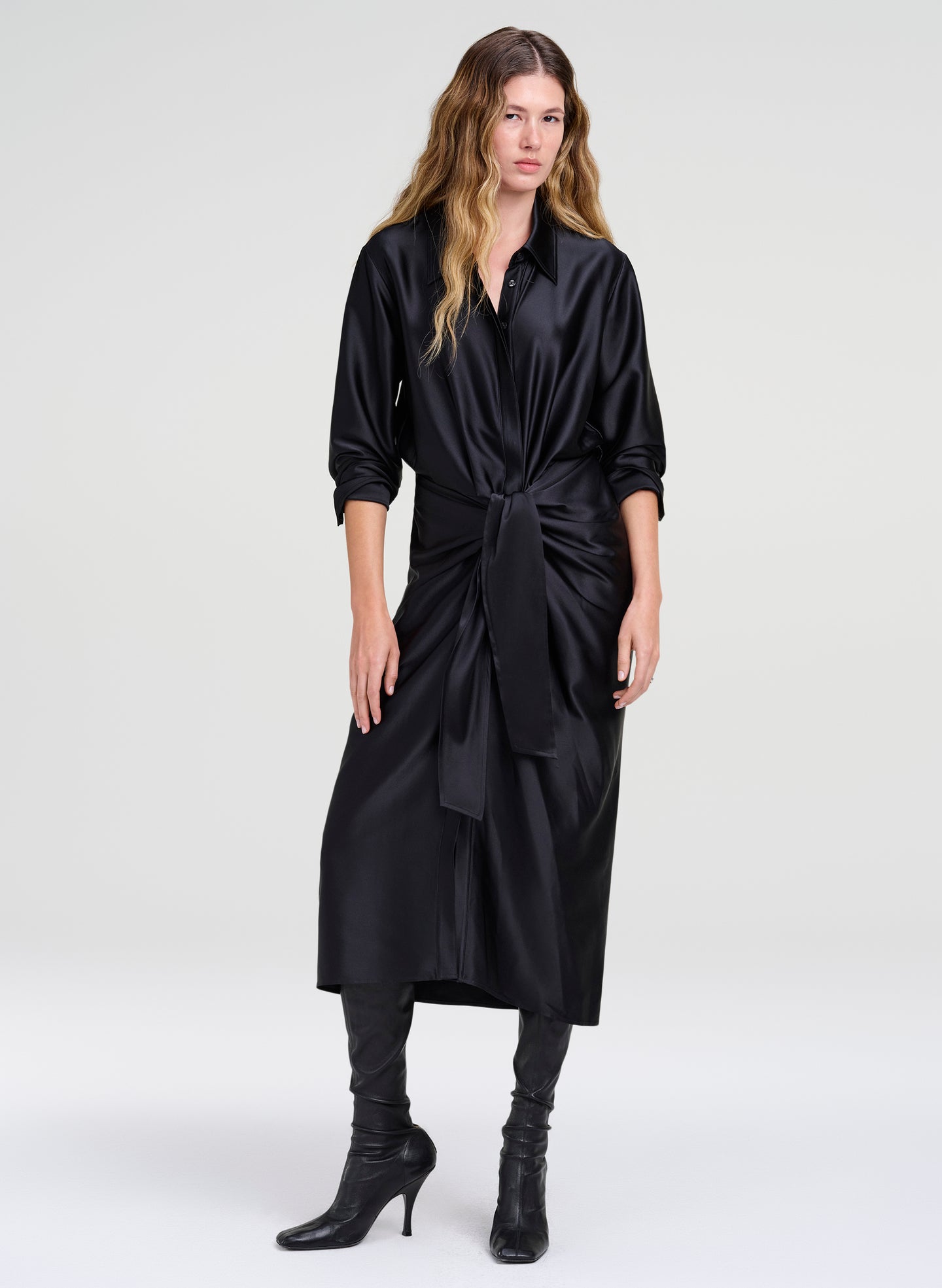 A woman modeling a black satin midi shirt dress with pushed up long sleeves and a self-tie waist belt, paired with black boots, against a light gray studio background.