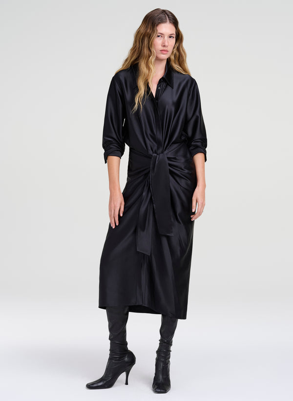 A woman modeling a black satin midi shirt dress with pushed up long sleeves and a self-tie waist belt, paired with black boots, against a light gray studio background.