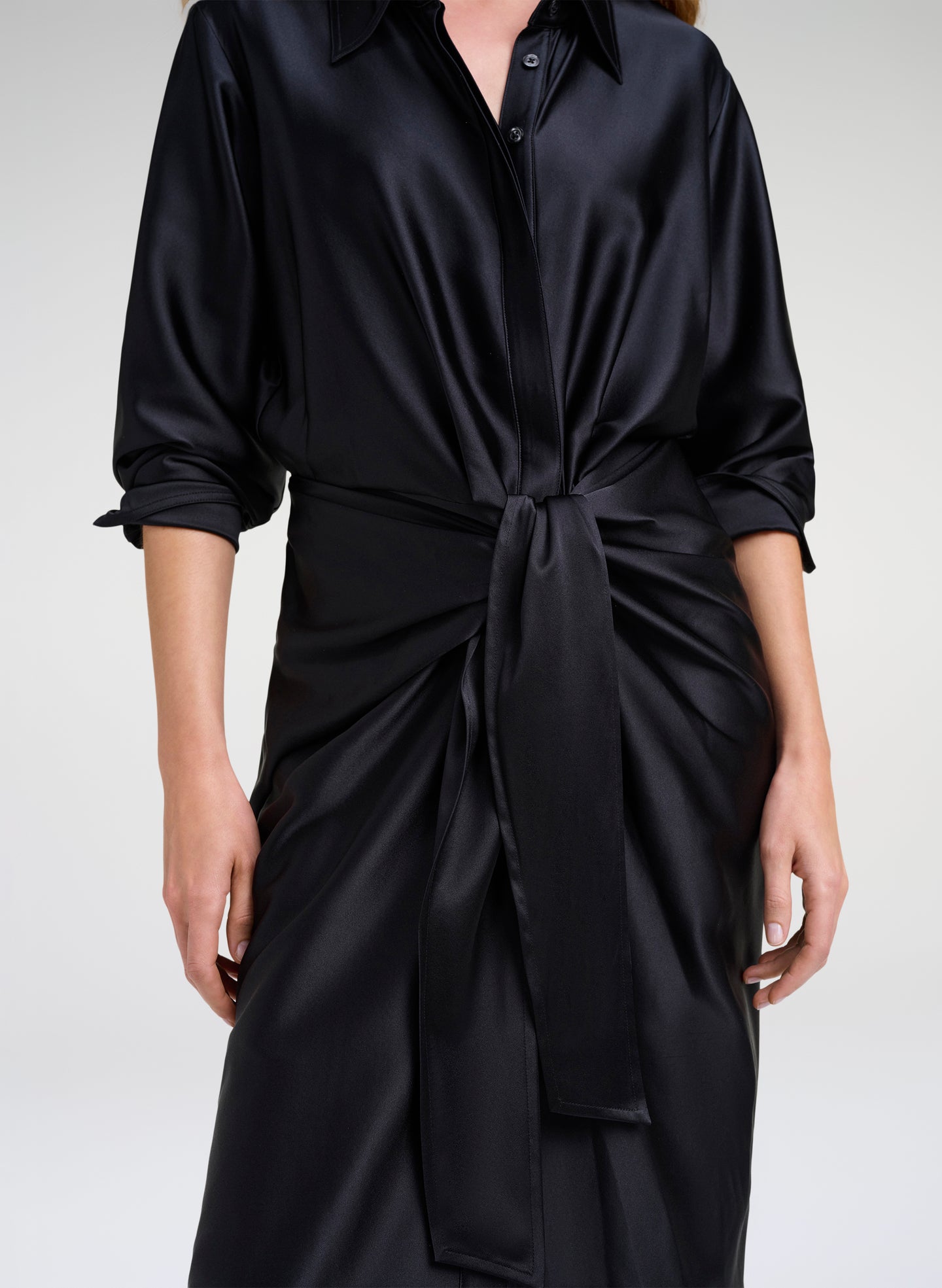 A close-up of a model wearing a black satin shirt dress with rolled-up sleeves and a self-tie belt at the waist, set against a plain light gray studio background.