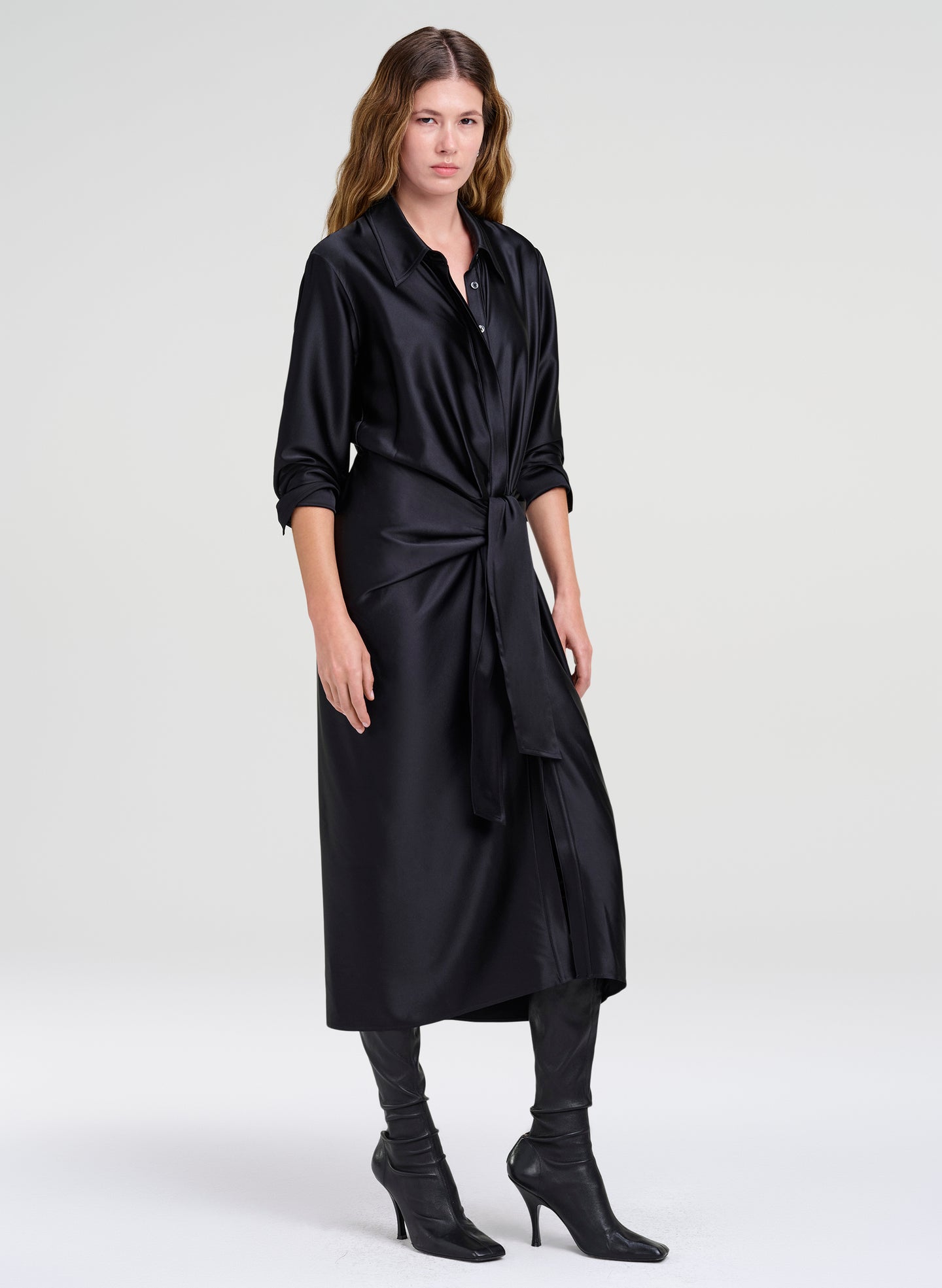 A woman modeling a black satin-like long shirt dress with a shawl collar, button front, rolled sleeves, and a self-tie waist, paired with black leggings and black heeled ankle boots against a light gray studio background.