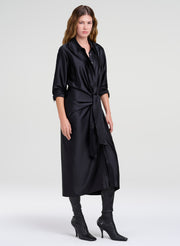 A woman modeling a black satin-like long shirt dress with a shawl collar, button front, rolled sleeves, and a self-tie waist, paired with black leggings and black heeled ankle boots against a light gray studio background.