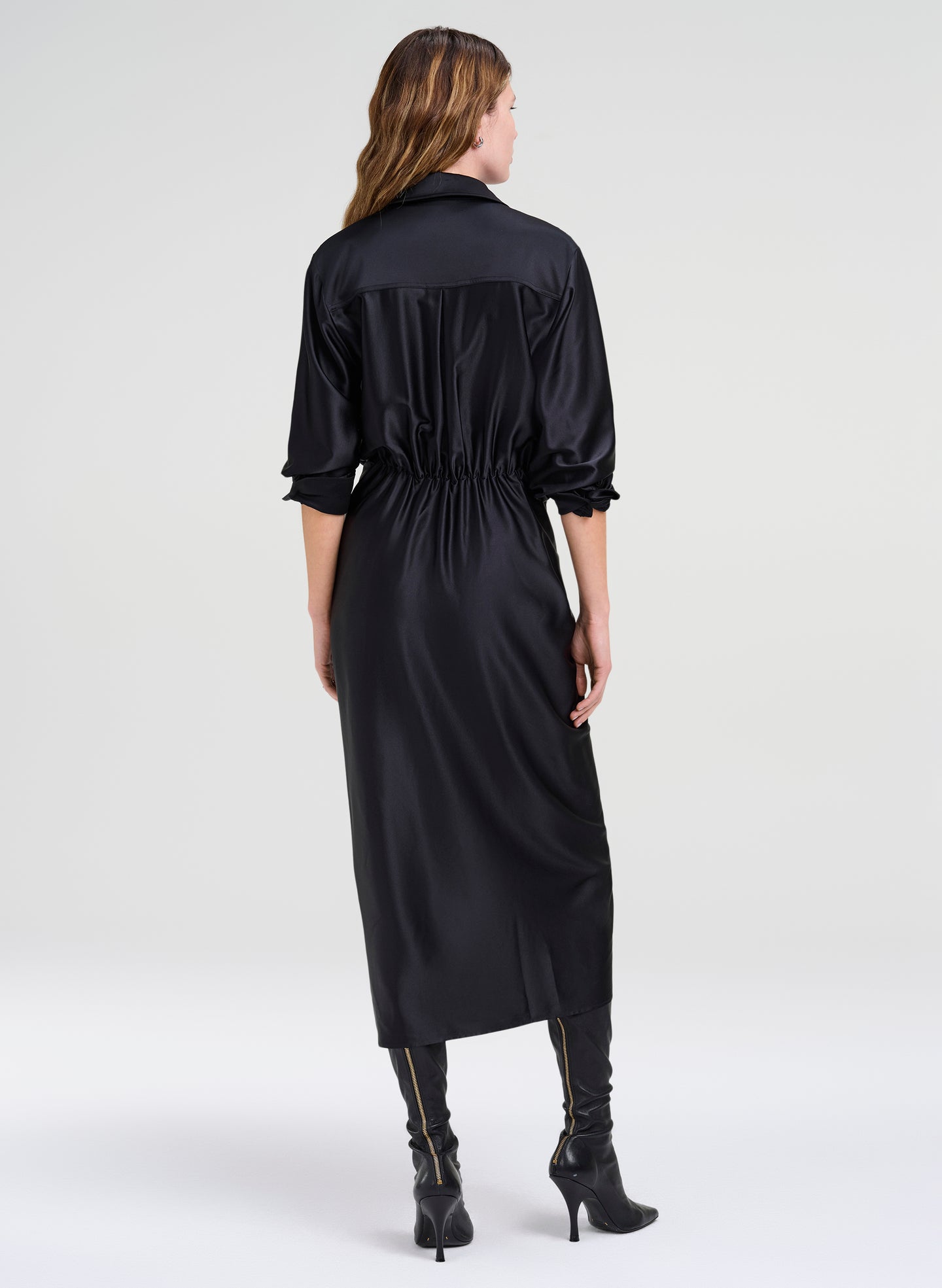 A woman viewed from the back wearing a black satin midi shirt dress with three-quarter sleeves and a gathered waist, paired with black heeled boots, against a light gray studio background.