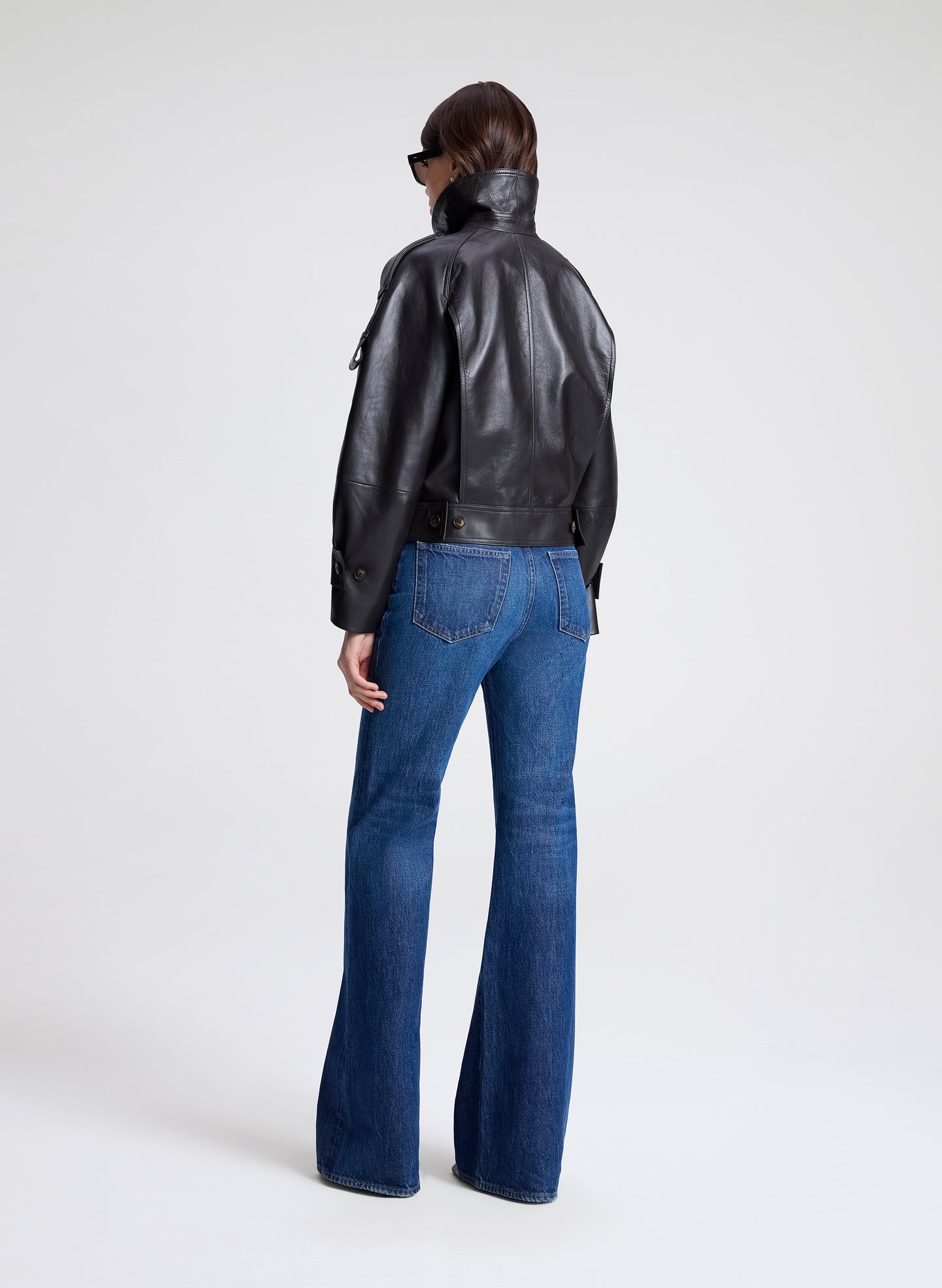 Woman wearing a dark brown leather jacket, black shirt, and blue jeans on a light gray background