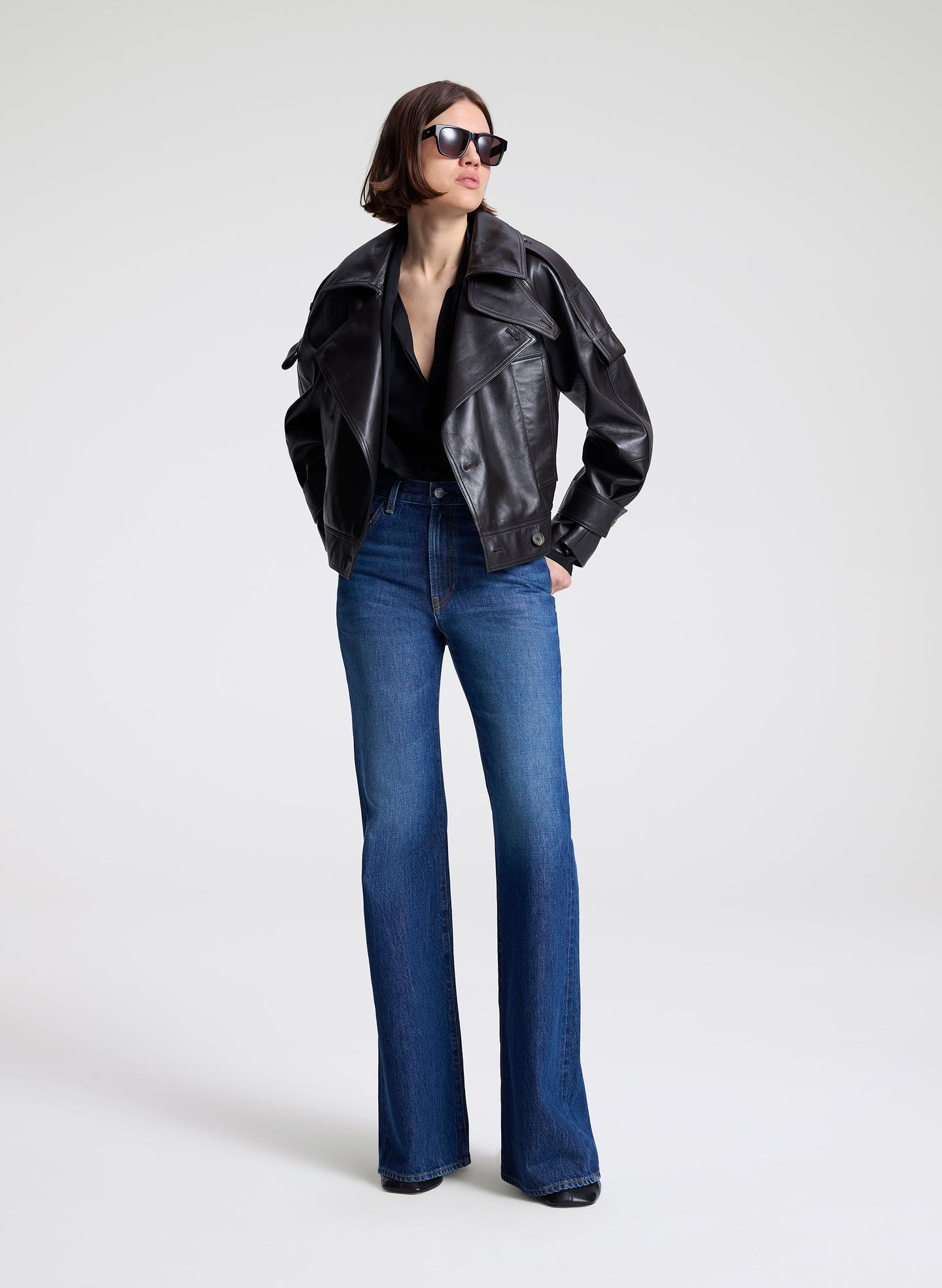 Woman wearing a dark brown leather jacket, black shirt, and blue jeans on a light gray background