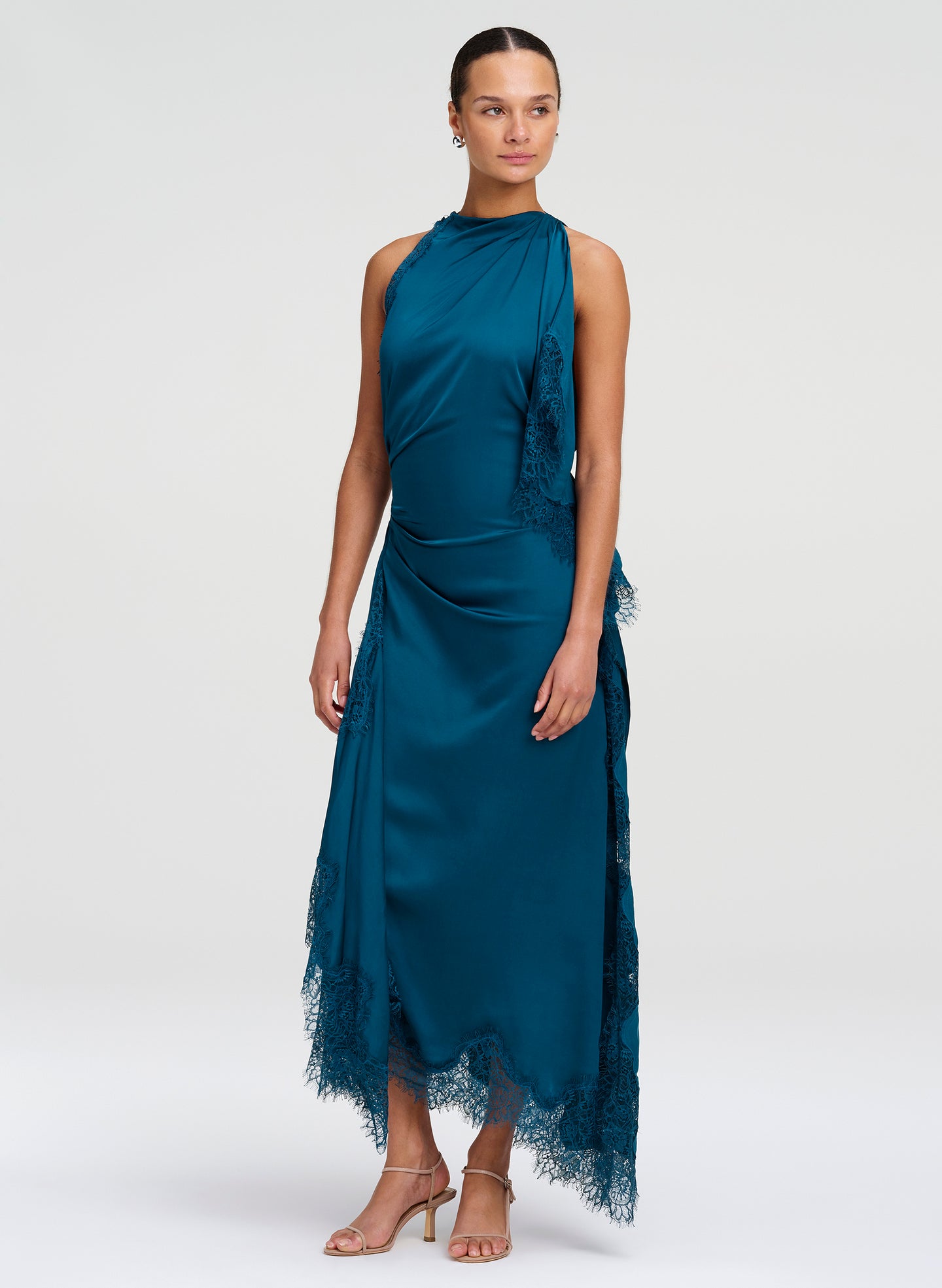 A model wearing a sleeveless, high-neck teal blue satin midi dress with lace ruffle details and a sheer lace hem, paired with tan high-heeled sandals, against a light gray studio background.