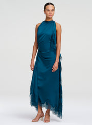 A model wearing a sleeveless, high-neck teal blue satin midi dress with lace ruffle details and a sheer lace hem, paired with tan high-heeled sandals, against a light gray studio background.